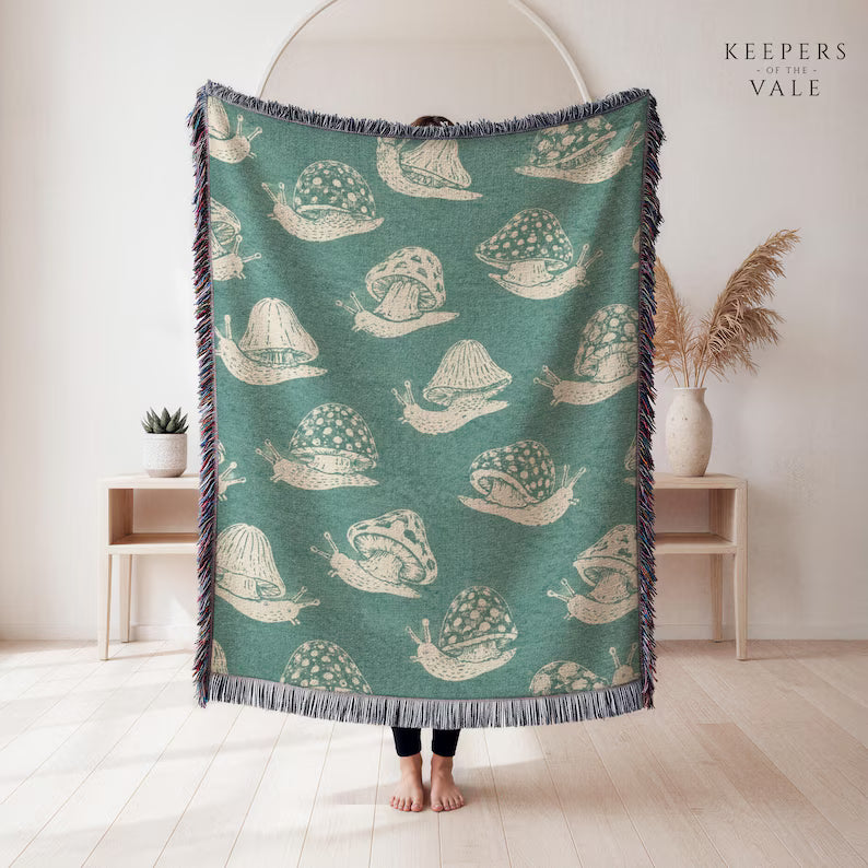 A person holds up a large woven throw blanket in a bright, minimalist room. The blanket has a teal green background with a repeating cream-colored snail pattern and dark blue-gray fringe on all sides.