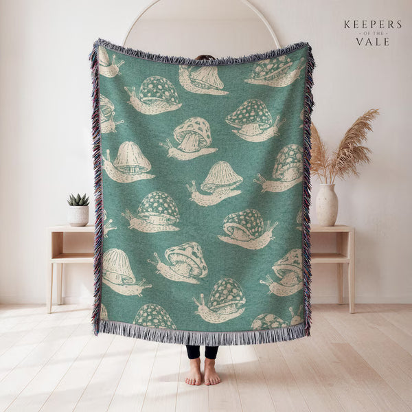 A person holds up a large woven throw blanket in a bright, minimalist room. The blanket has a teal green background with a repeating cream-colored snail pattern and dark blue-gray fringe on all sides.
