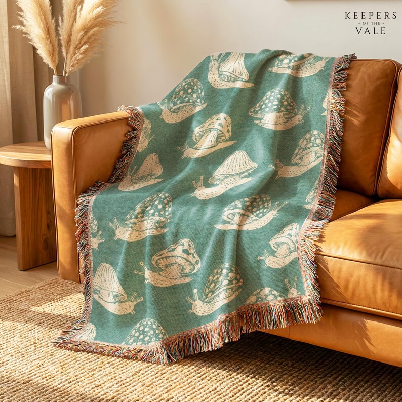 A teal throw blanket with a cream snail pattern and multicolor fringe, draped over a tan leather armchair in a warm, sunlit room. The brand name 'KEEPERS FOR LIFE VALE' is printed in the top right corner.